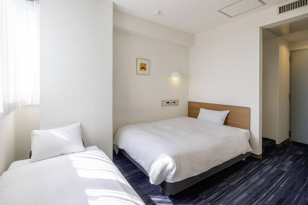 A hotel room with two single beds, a small bedside table, a wall-mounted light, and a picture on the wall.