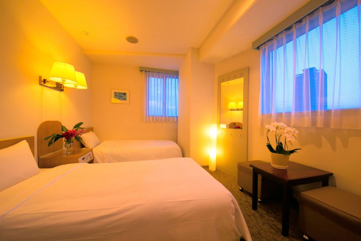 A hotel room with two beds, a bedside table with a lamp, a small table with a vase of flowers, a window with curtains, and a mirror.