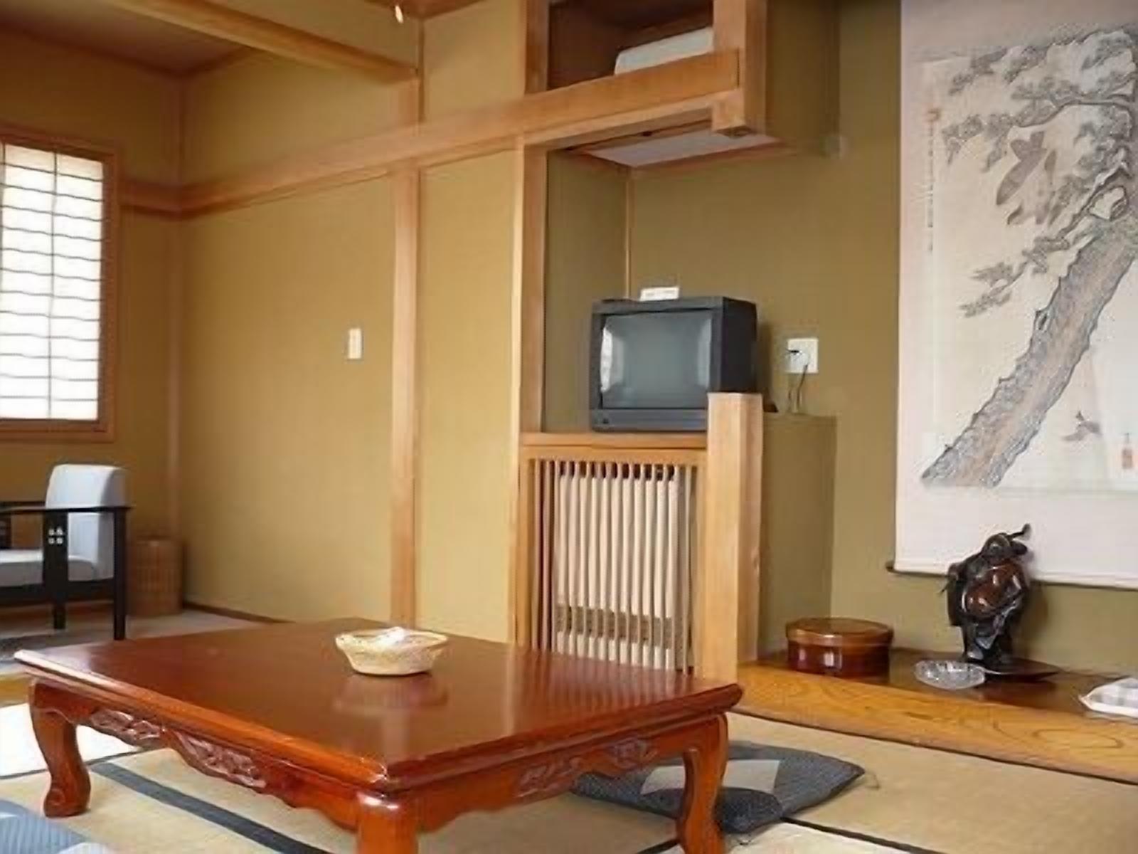 Ein traditioneller japanischer Raum mit Tatami-Böden, einem niedrigen Holztisch, einem Fernseher auf einem Schrank und einem Bild an der Wand.