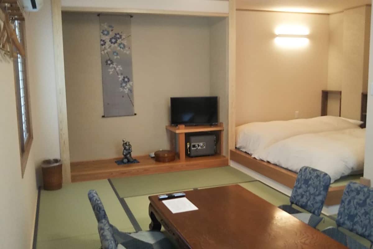Una habitación pequeña con una cama, un televisor sobre un soporte, una mesa con sillas y un adorno colgante.