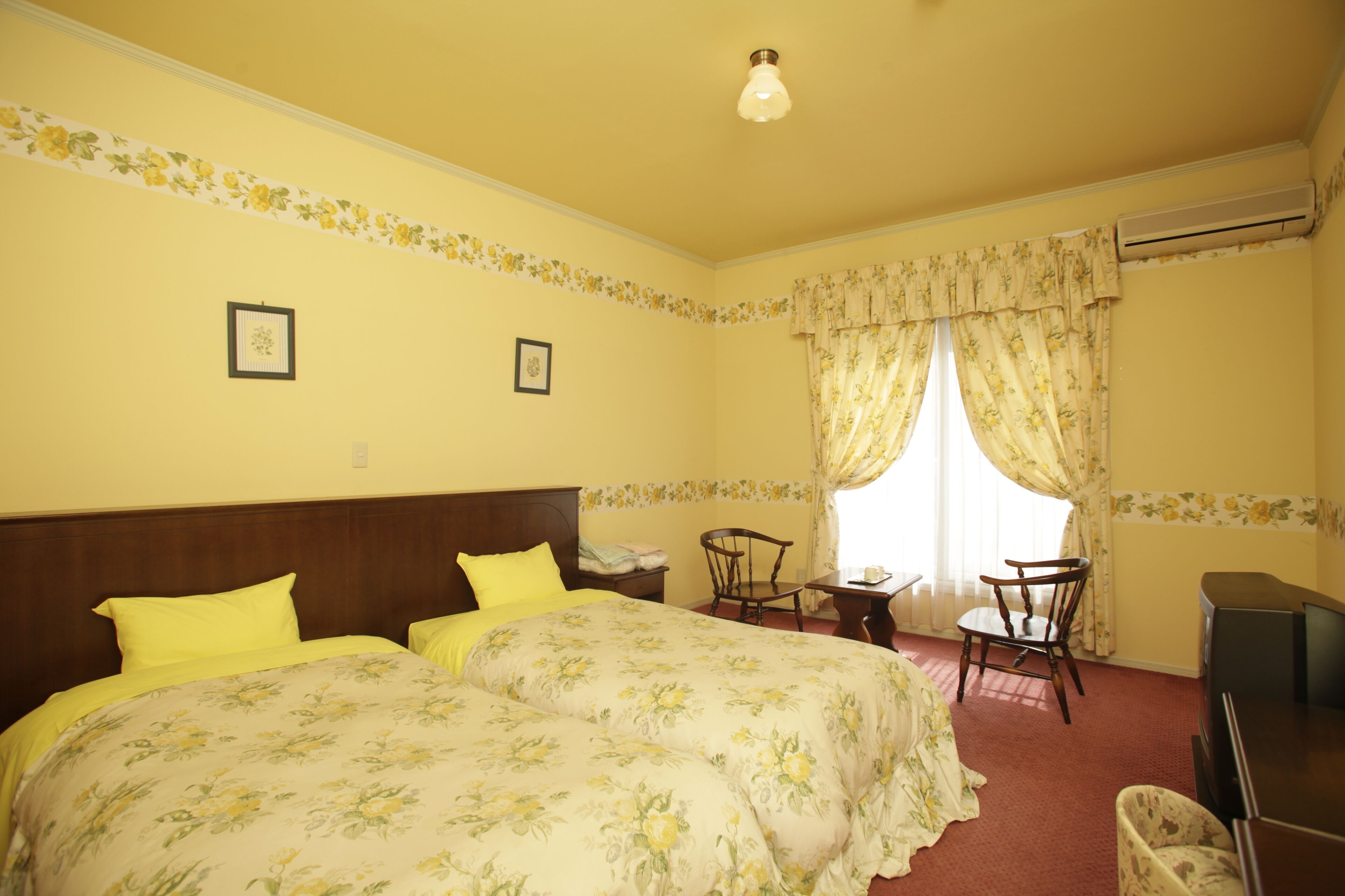A hotel room with two beds, a desk, a chair, an air conditioning unit, and a window with curtains.