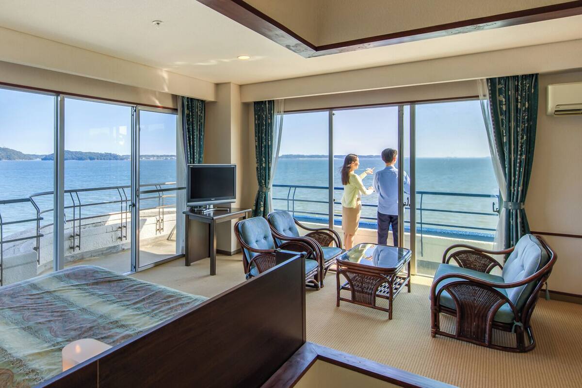 A room with a bed, a TV, a table, and chairs, with a view of the sea through large windows.