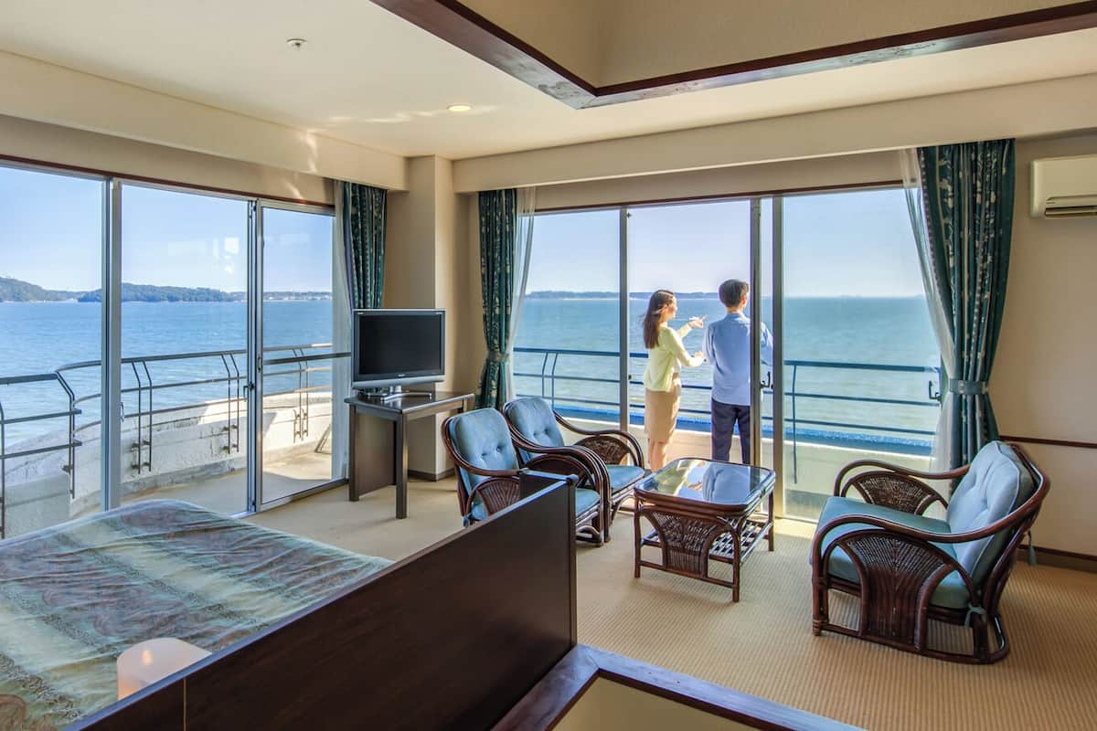A room with a bed, a TV, a table, and chairs, with a view of the sea through large windows.