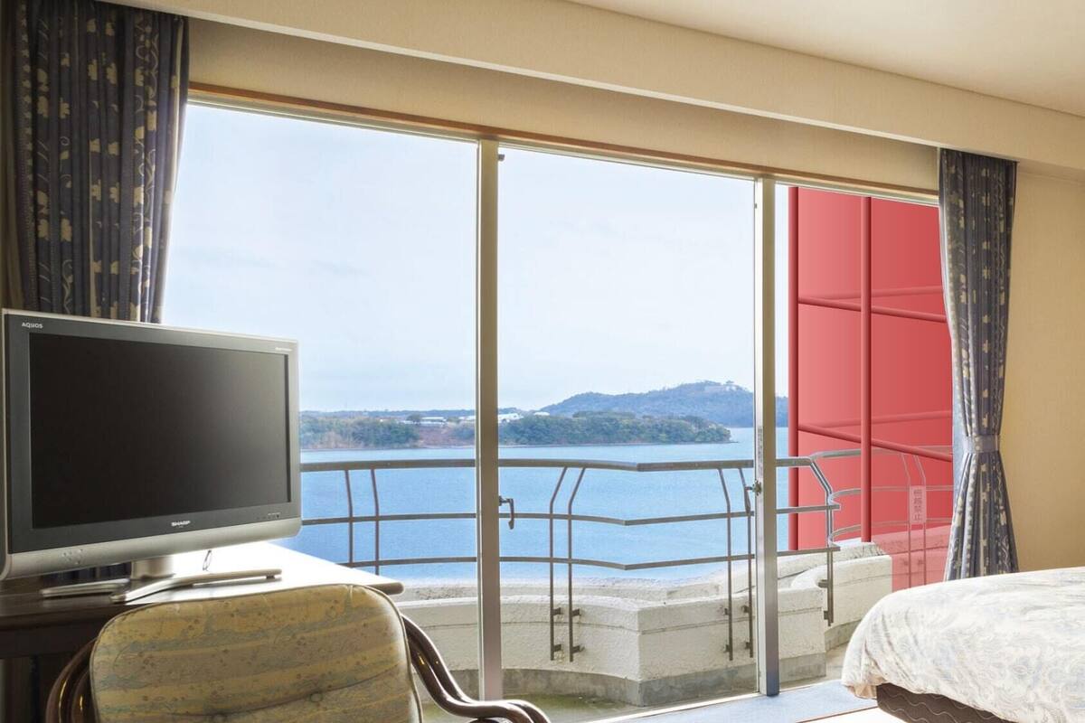 A hotel room with a television, a bed, a balcony with a view of a body of water and hills, and an air conditioning unit.