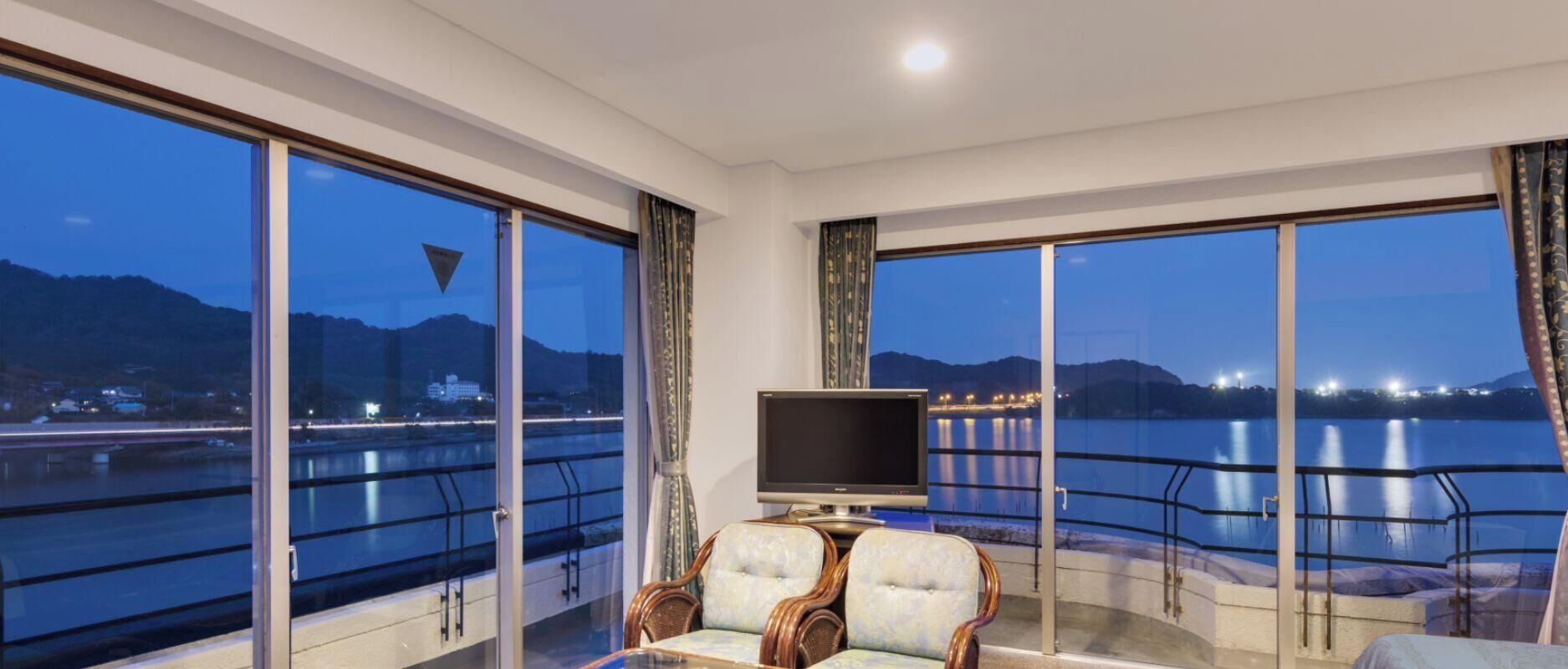 A room with large windows, a television, and a seating area with a view of a body of water and mountains.