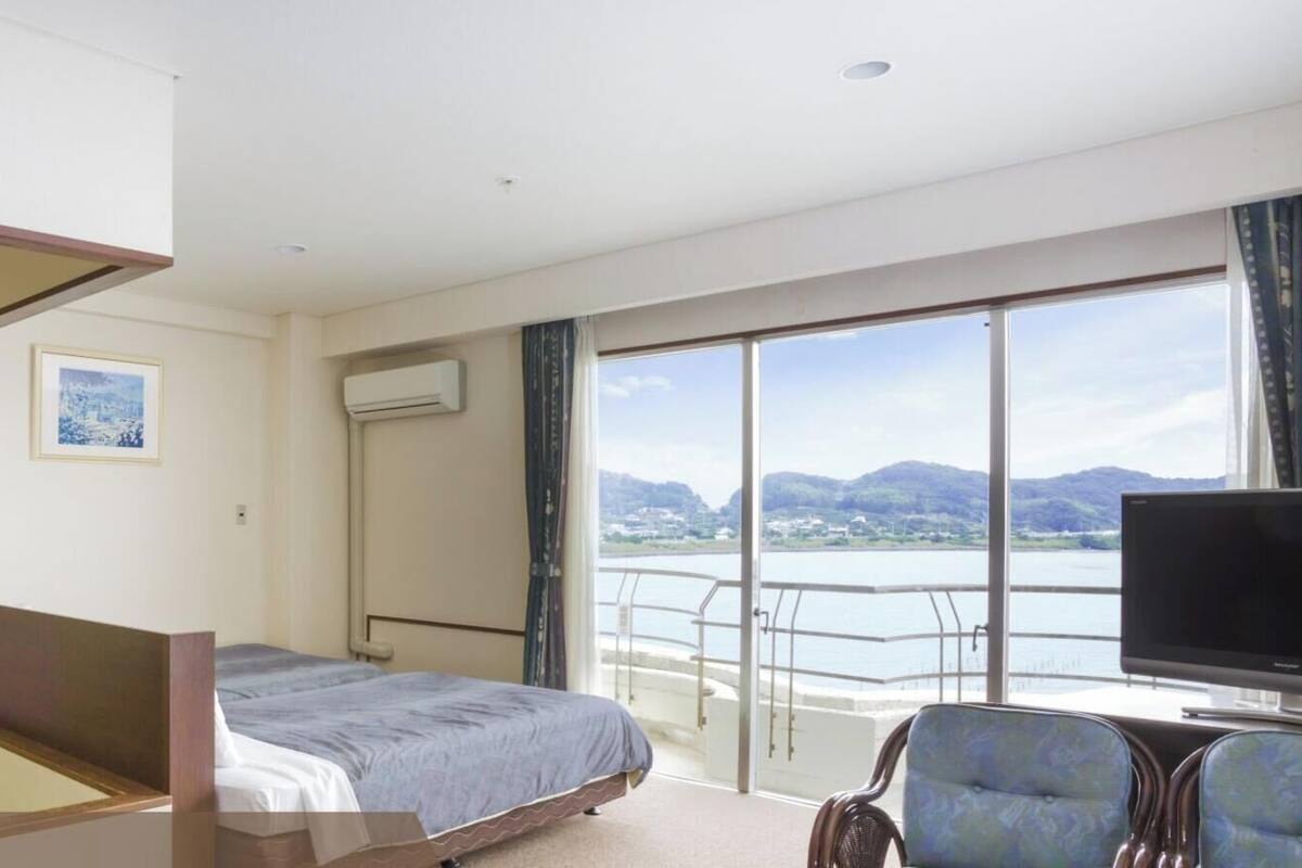 A hotel room with a bed, a television, a chair, a picture on the wall, and a balcony with a view of the water.