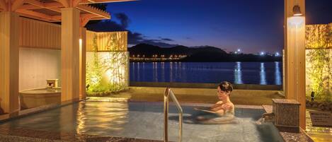 A person sitting in a hot spring pool with a view of a body of water and mountains at dusk.