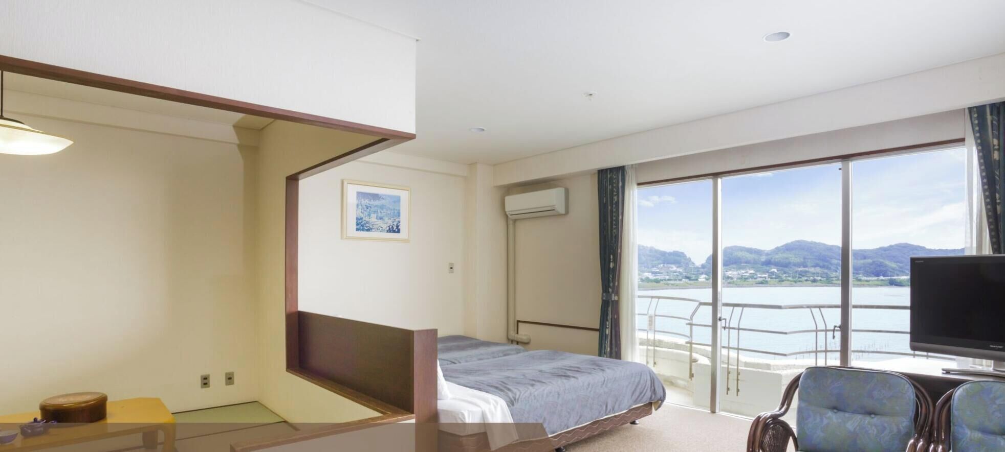 A hotel room with a bed, a television, a chair, a picture on the wall, and a balcony with a view of the water.