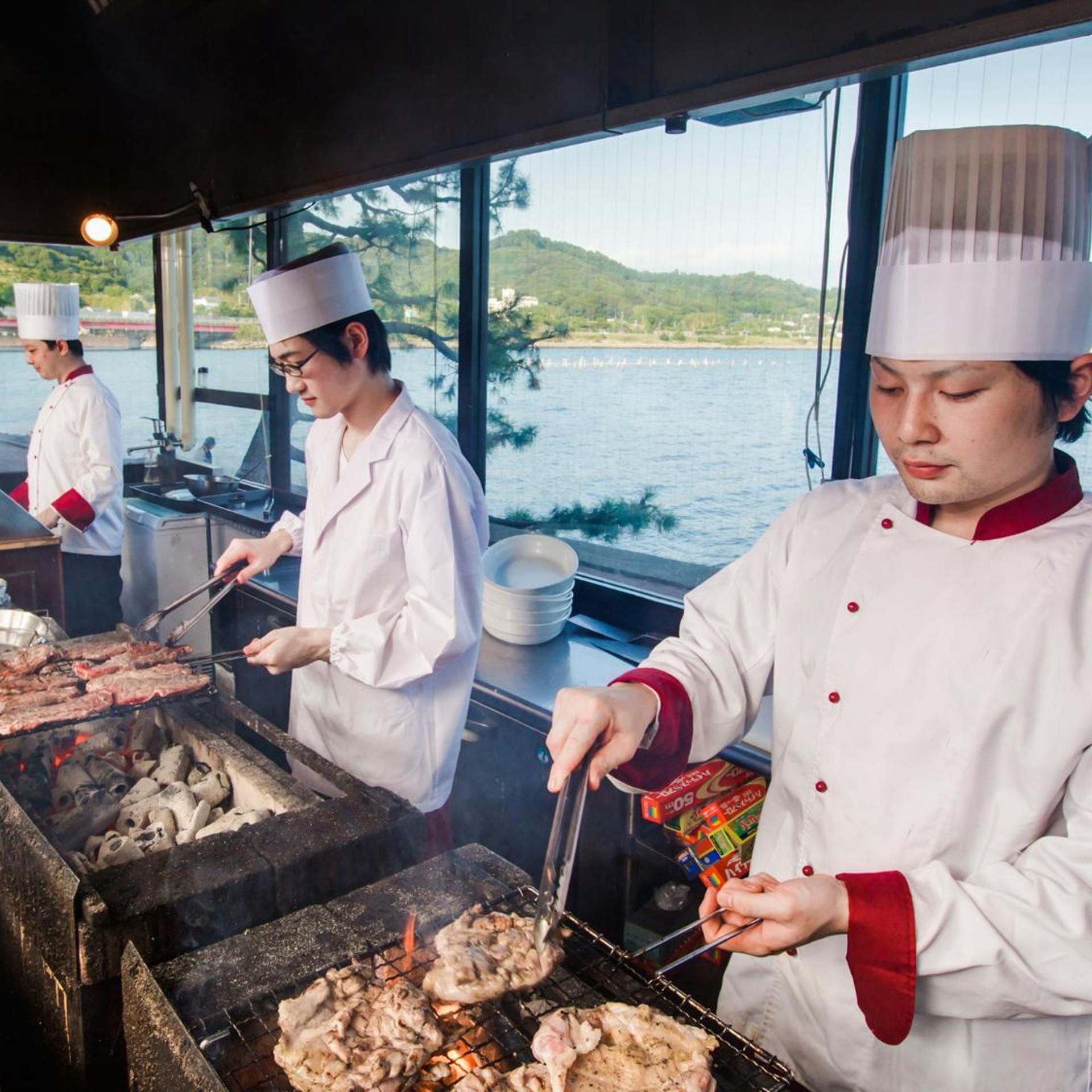Köche grillen Fleisch in einer Außenküche mit Blick auf ein Gewässer und Hügel im Hintergrund.
