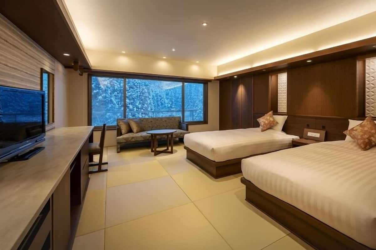 A modern hotel room with two beds, a sofa, a TV, and large windows with a view of snow-covered trees.