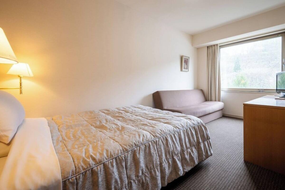 A hotel room with a bed, a desk, a chair, a window, and a lamp.