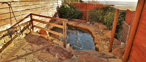 Una escalera de piedra conduce a un área de aguas termales, con una valla de madera y vista al océano.