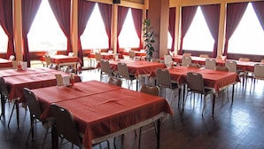 A dining area with multiple tables and chairs, large windows with curtains, and a potted plant. - Hotel Innoshima (Onomichi)