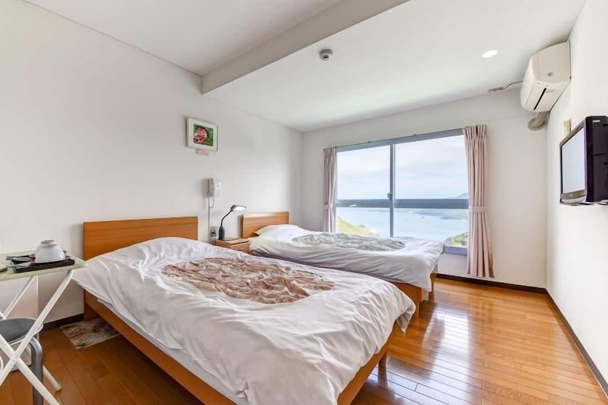 A hotel room with two beds, a desk with a kettle, a bedside table, a window with a view, and an air conditioning unit.
