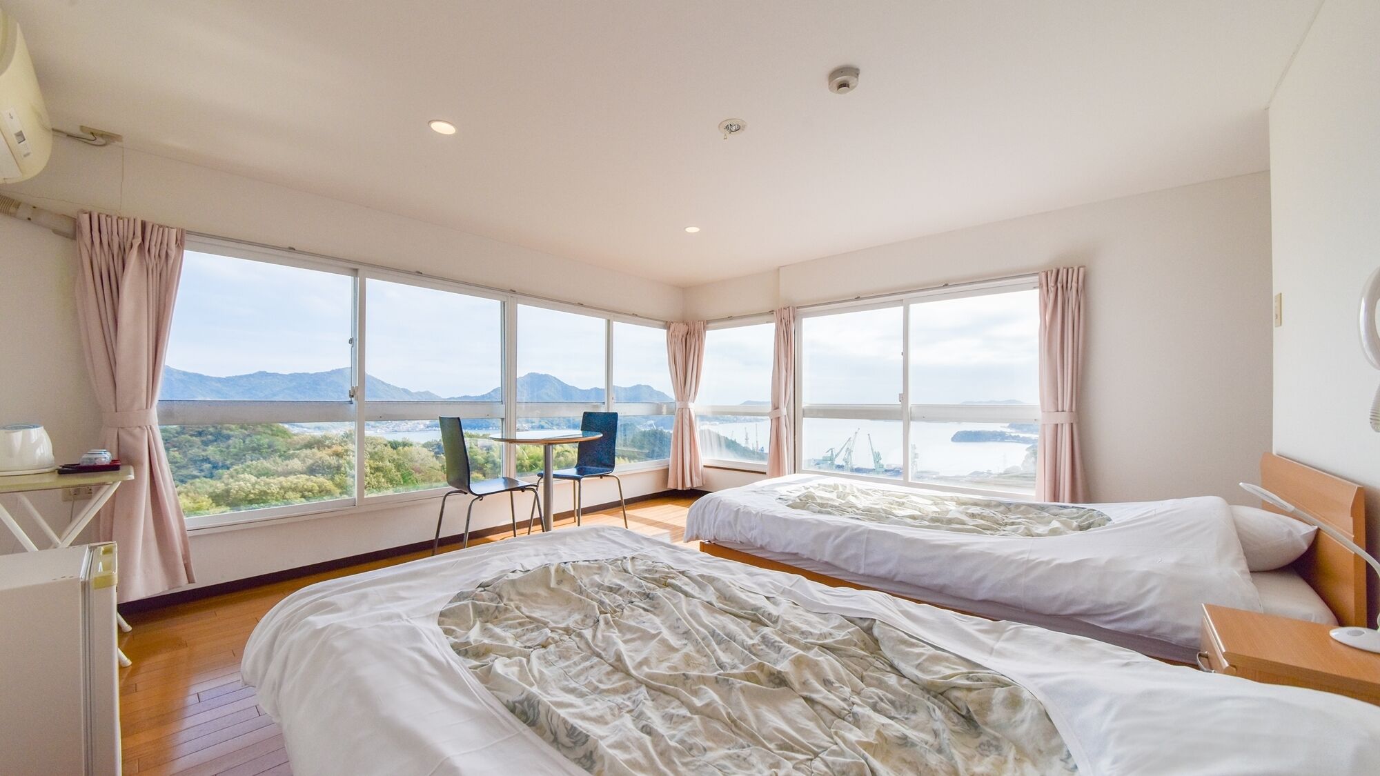 A hotel room with two beds, a desk, a chair, and large windows offering a view of a landscape with mountains and a body of water.