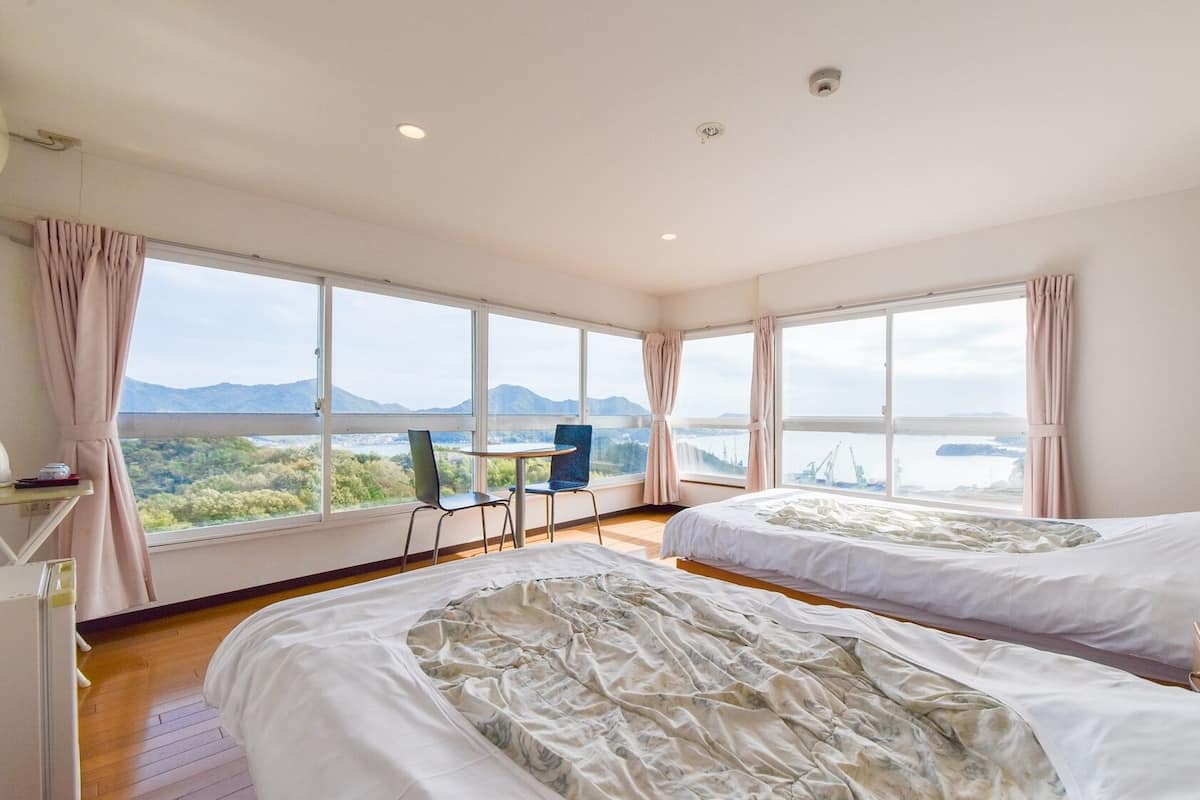 A hotel room with two beds, a desk, a chair, and large windows offering a view of a landscape with mountains and a body of water.