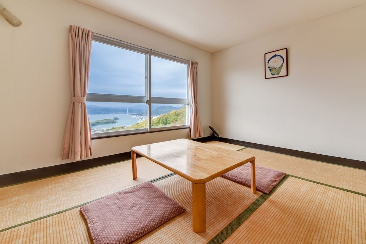 A room with tatami flooring, a wooden table, and a view of a body of water and mountains.