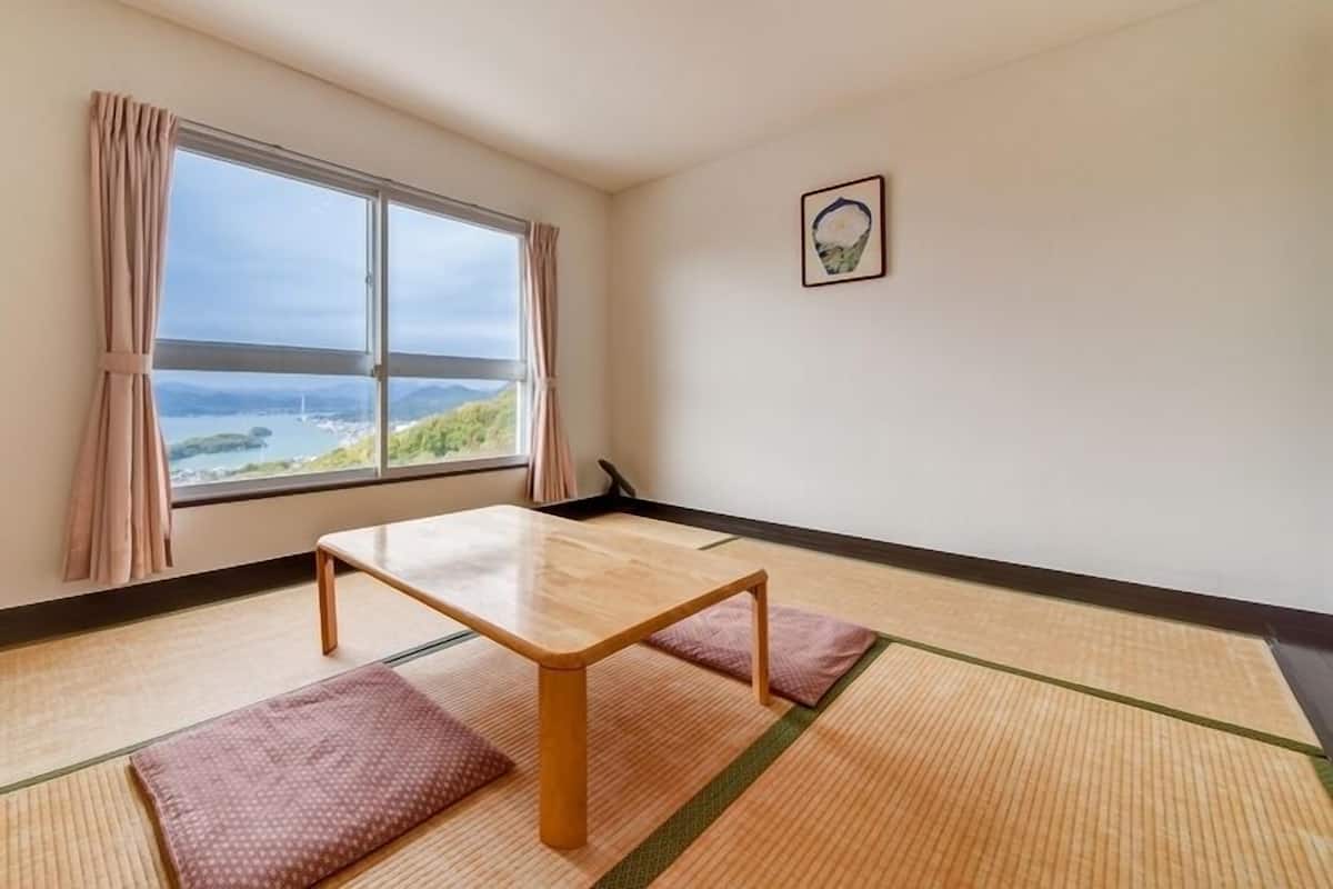 A room with tatami flooring, a wooden table, and a view of a coastal landscape through a large window.