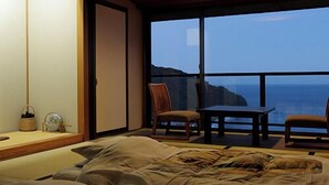 A bedroom with a bed, a small table, two chairs, and a large window offering a view of the sea. - Atagawa Onsen Suiko (Higashiizu)