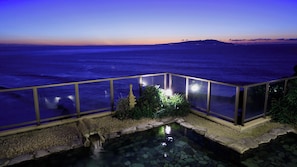 Vue nocturne d’une zone côtière avec une piscine, une balustrade en verre et un chemin de pierre menant à l’eau.