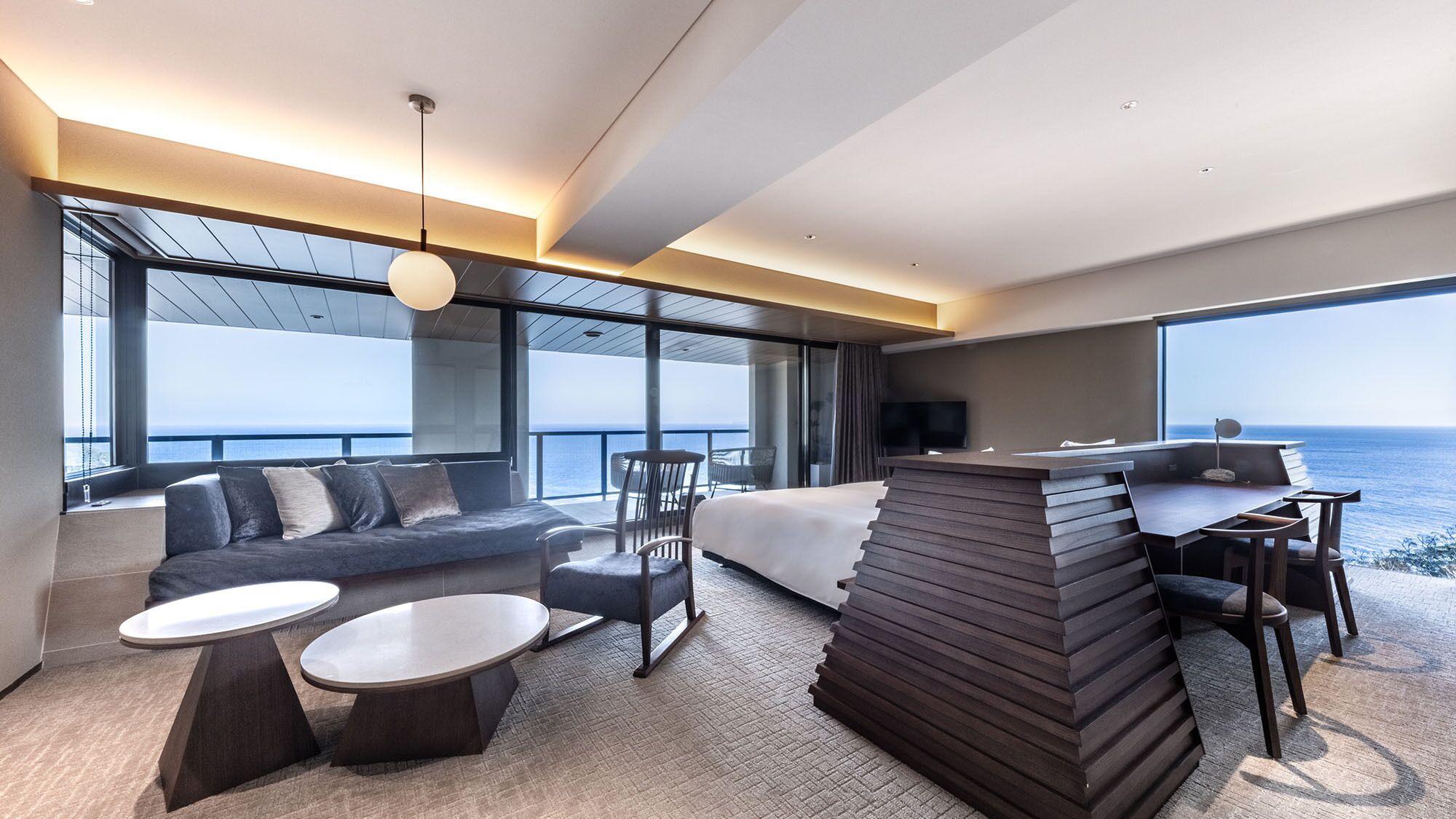 Una habitación de hotel moderna con una cama grande, un área de descanso con sofá y sillones, una mesa de comedor y vistas al océano.