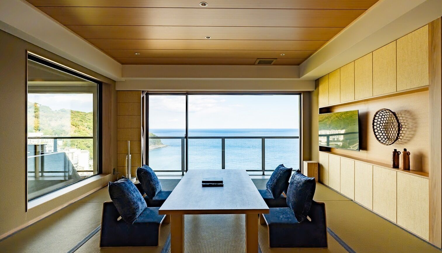 Une pièce moderne dotée d’une grande table, de sièges confortables, d’une vue sur la mer et d’un téléviseur fixé au mur.