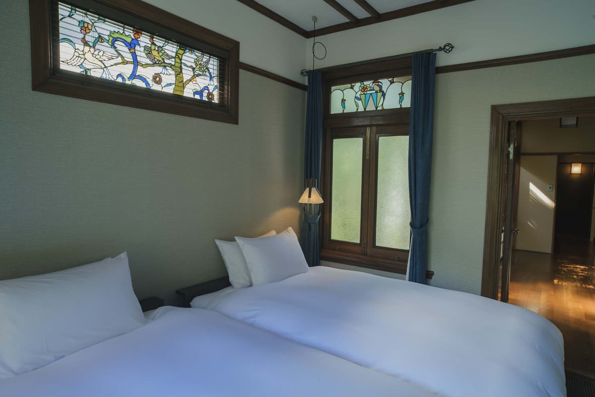 A hotel room with two beds, a large window with stained glass, and a door leading to another room.