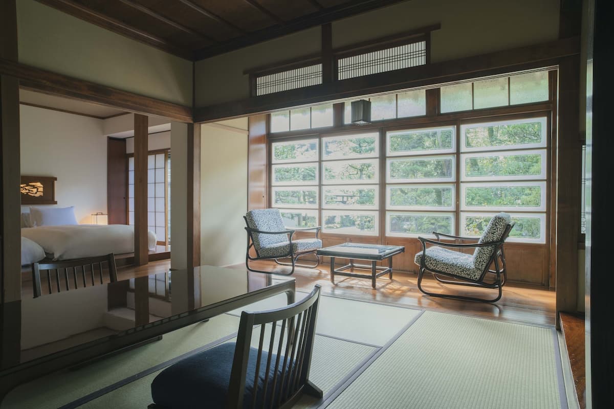 A traditional Japanese-style room with tatami flooring, a low table, two patterned chairs, and a view through large windows.