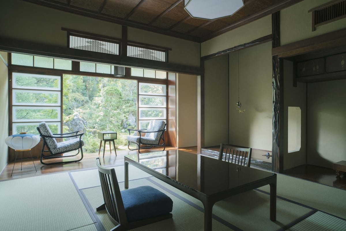 A traditional Japanese room with tatami flooring, a low table, two patterned chairs, and large windows offering a view of greenery.