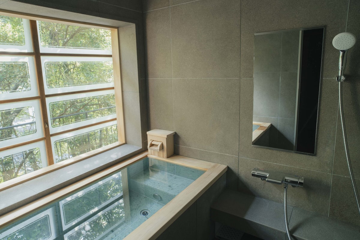 A modern bathroom with a glass-enclosed bathtub, a large window with a view of trees, and a wall-mounted shower.
