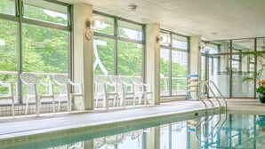 A swimming pool with clear glass walls, white plastic chairs, and a view of greenery outside.