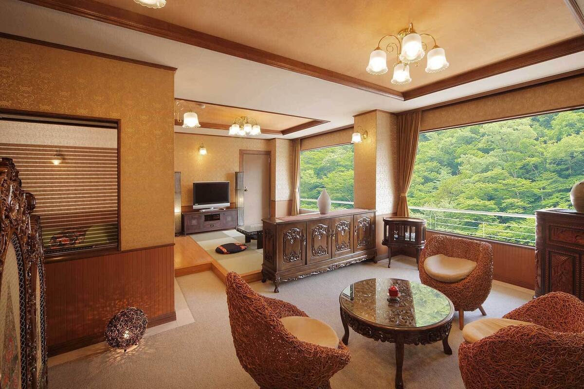 Amplia habitación con ventana grande que ofrece vistas al bosque, televisor, mueble y dos sillas de mimbre.