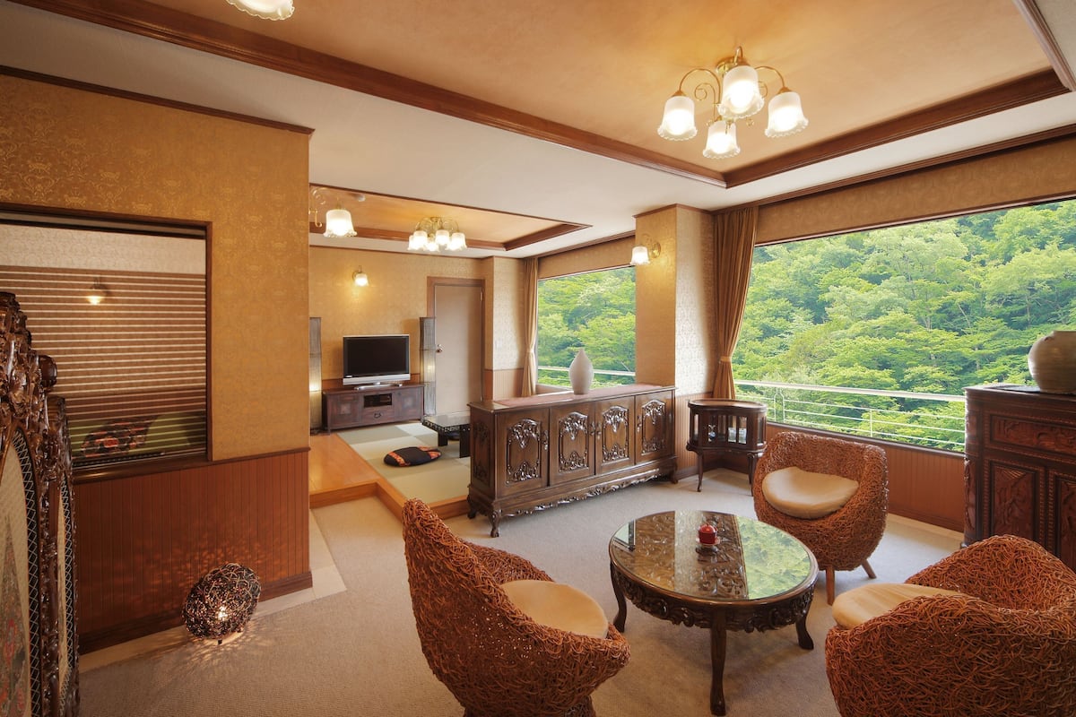 Una habitación tradicional japonesa con suelo de tatami, un bajo mueble de madera, un televisor y vistas a un bosque a través de un amplio ventanal.