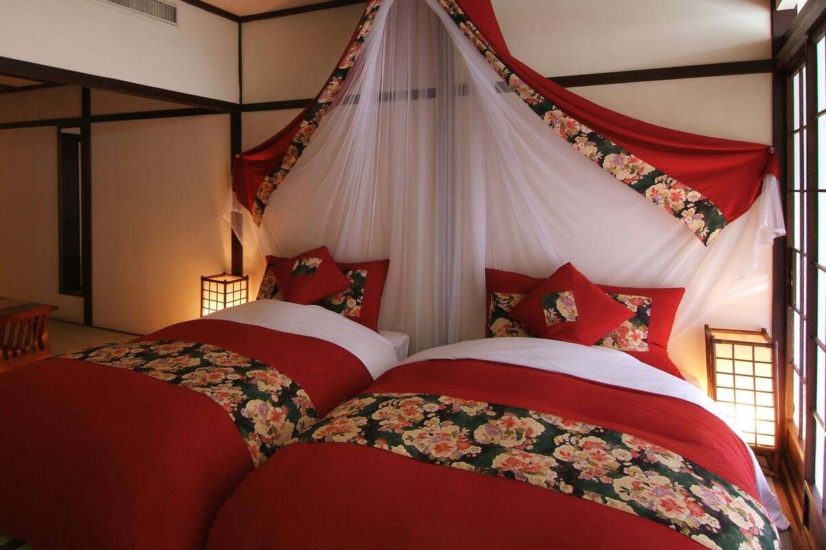 Un dormitorio tradicional japonés con cama a dosel, ropa de cama con estampado floral rojo y vistas a una habitación japonesa tradicional con suelo de tatami y una mesa baja.