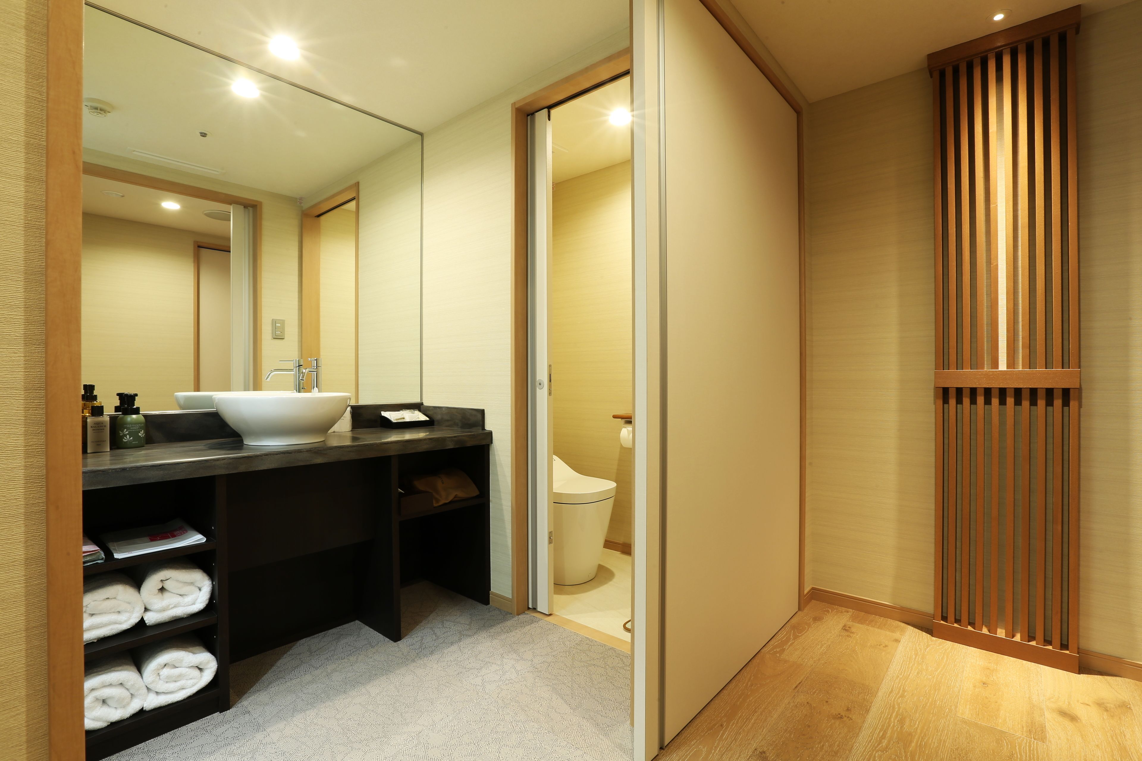 A modern bathroom with a large mirror, a sink, and a toilet. There are towels neatly folded on a shelf.