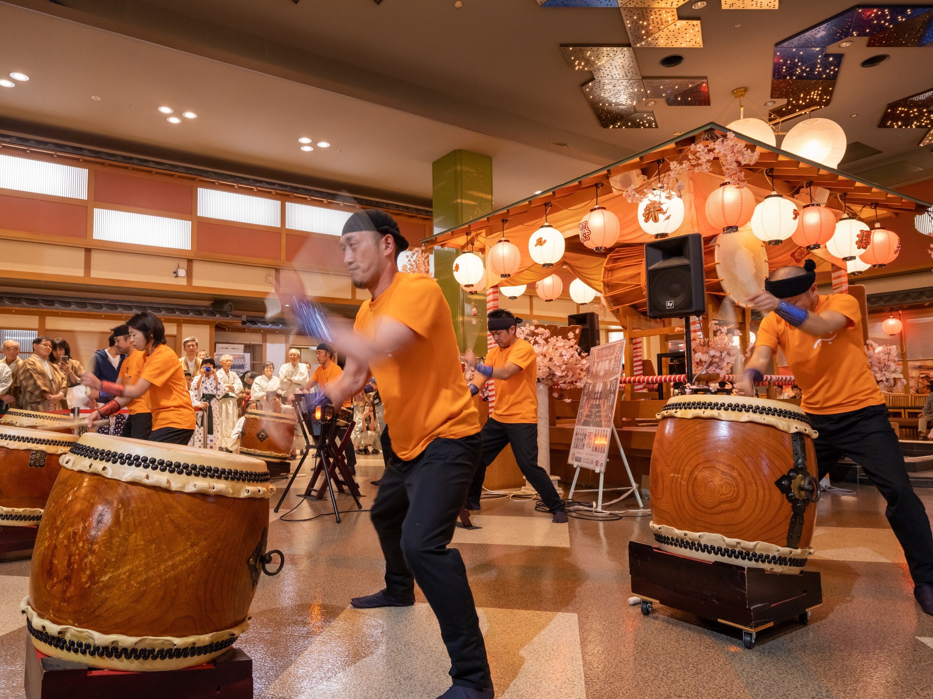 A group of people playing traditional drums in a spacious hall with a high ceiling and decorative lighting.