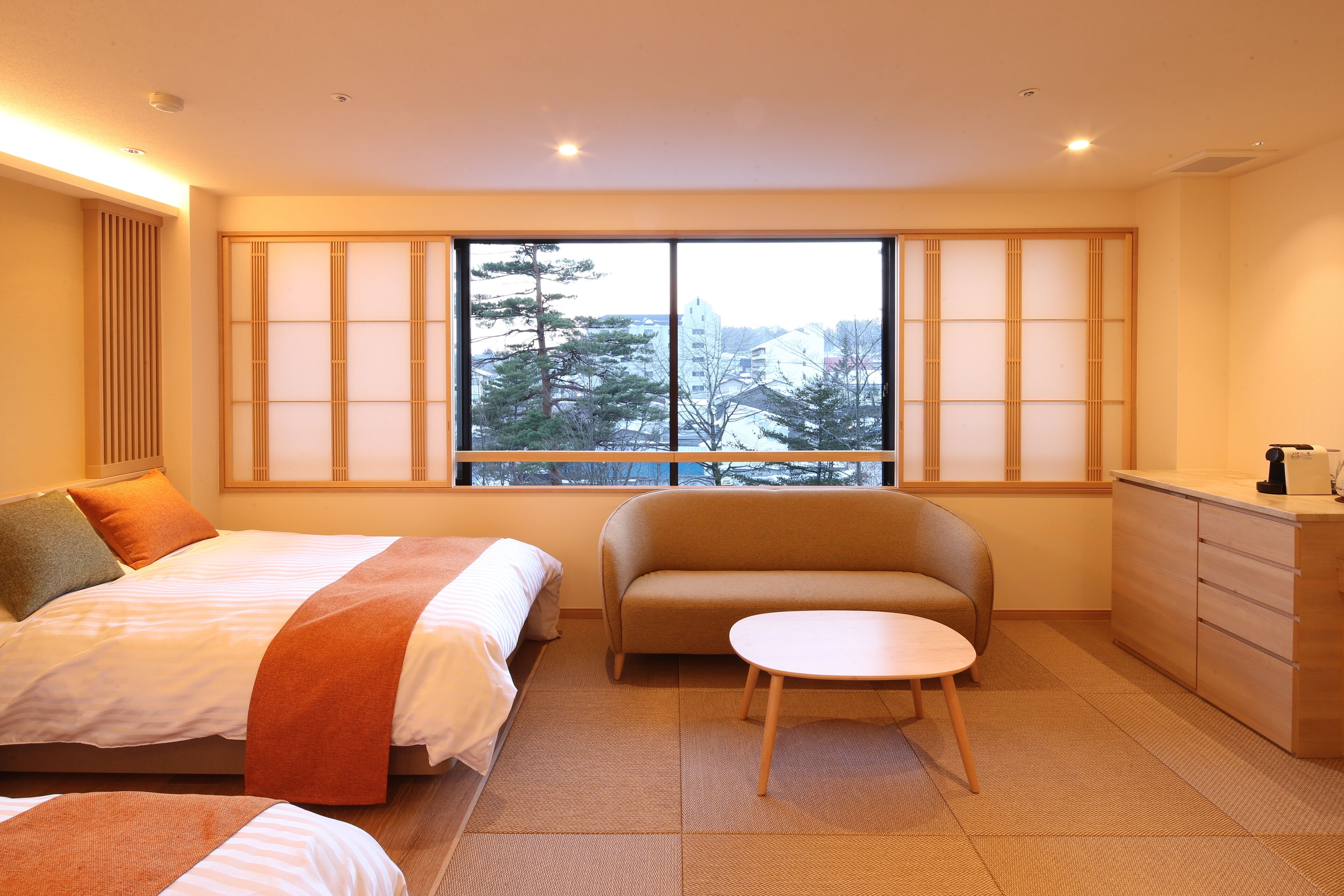 Ein Hotelzimmer mit Bett, Sofa, kleinem Tisch, Kommode und Fenster mit Blick auf Bäume und Gebäude.