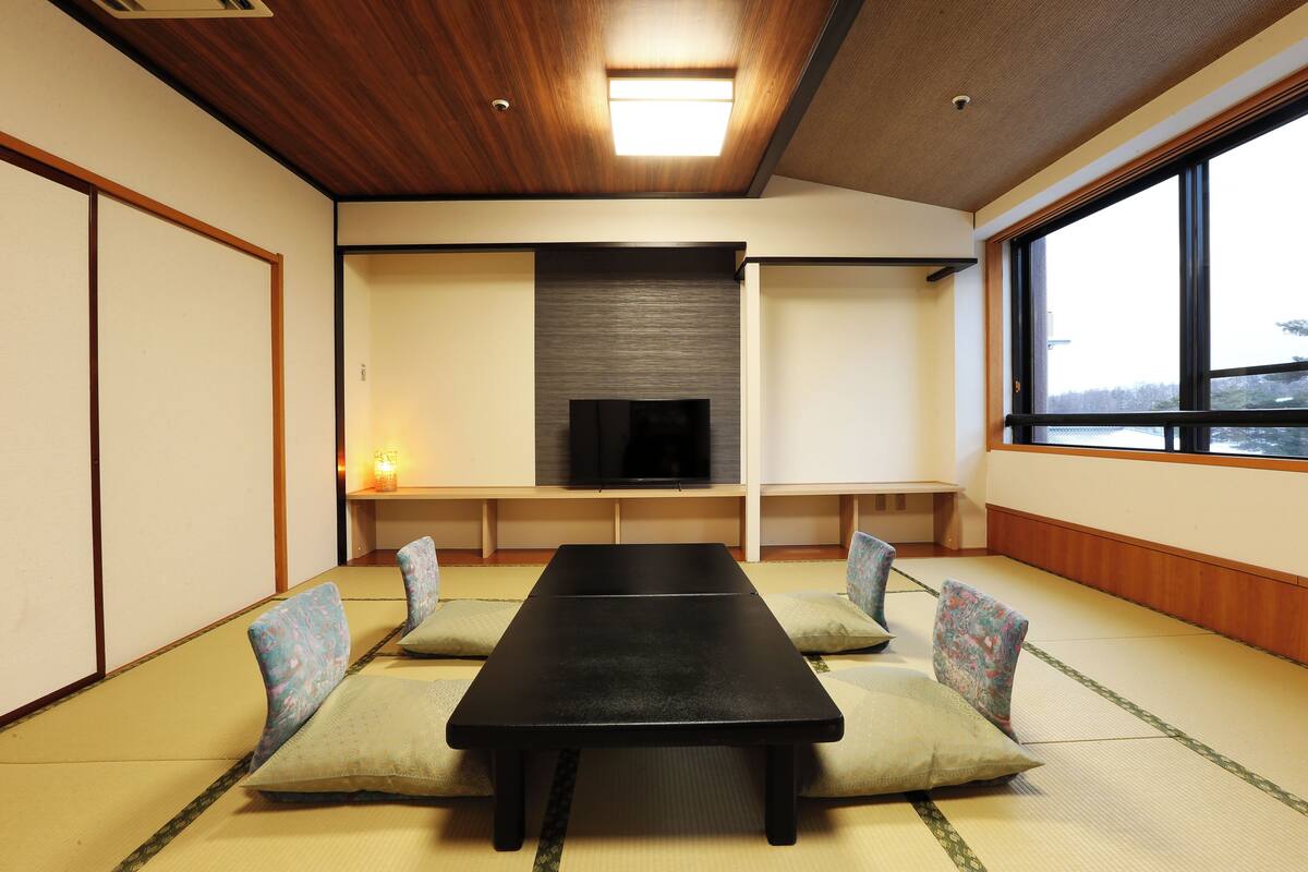 Une pièce avec un sol recouvert de tatamis, une table noire, des coussins, un téléviseur et une grande fenêtre.