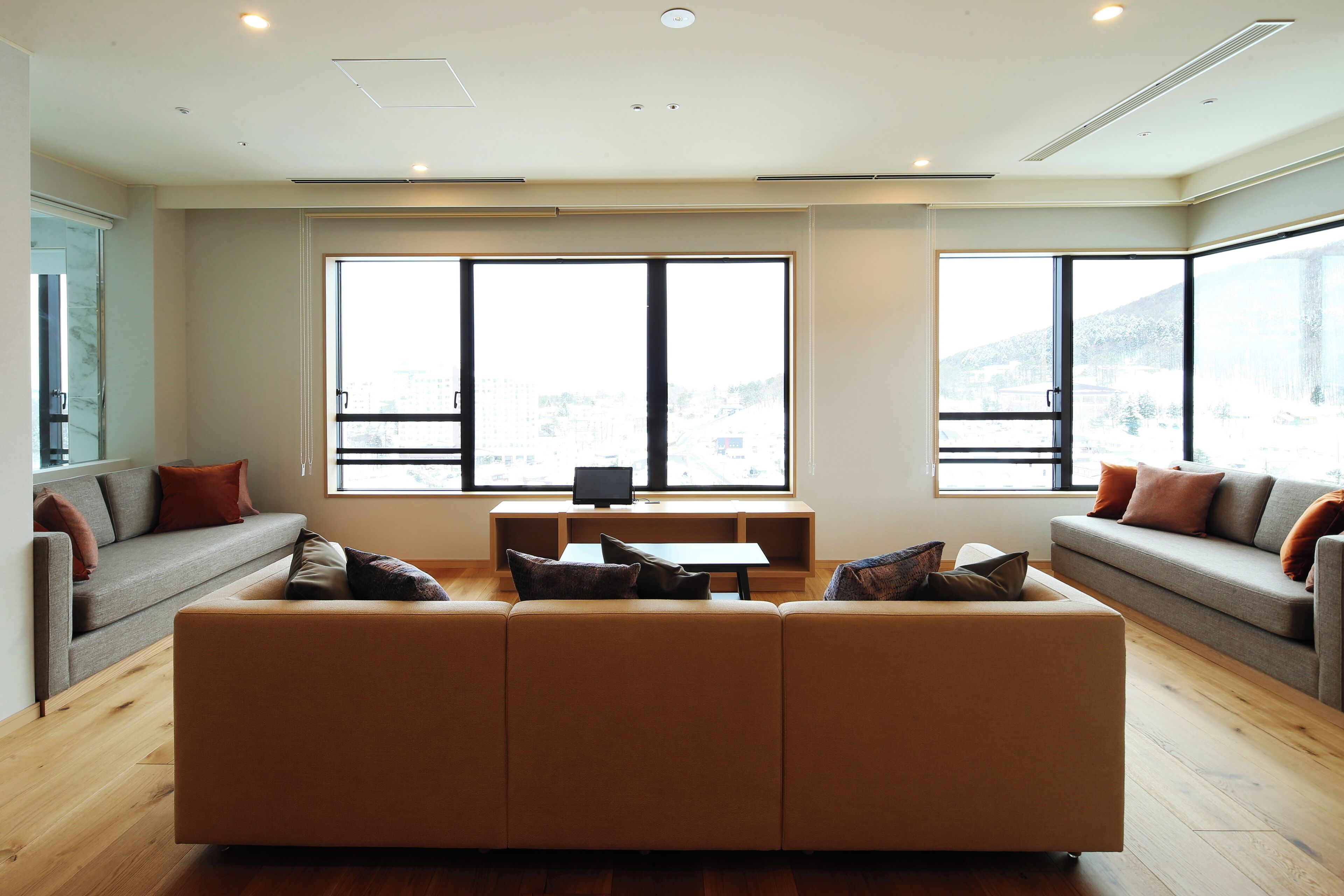 A modern living room with a large beige sectional sofa, a small table, and two couches with cushions. Large windows provide a view of the cityscape.
