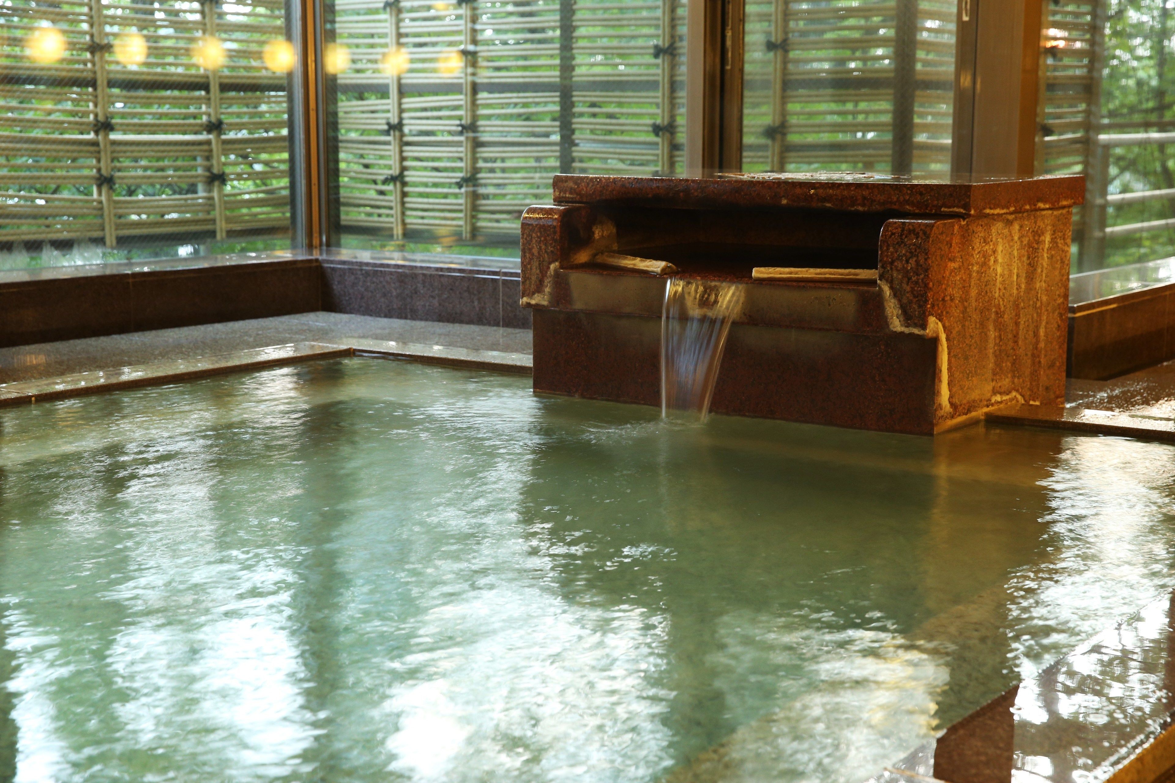 Una bañera de madera tradicional con una fuente de agua que fluye, frente a un fondo de bambú y una ventana.