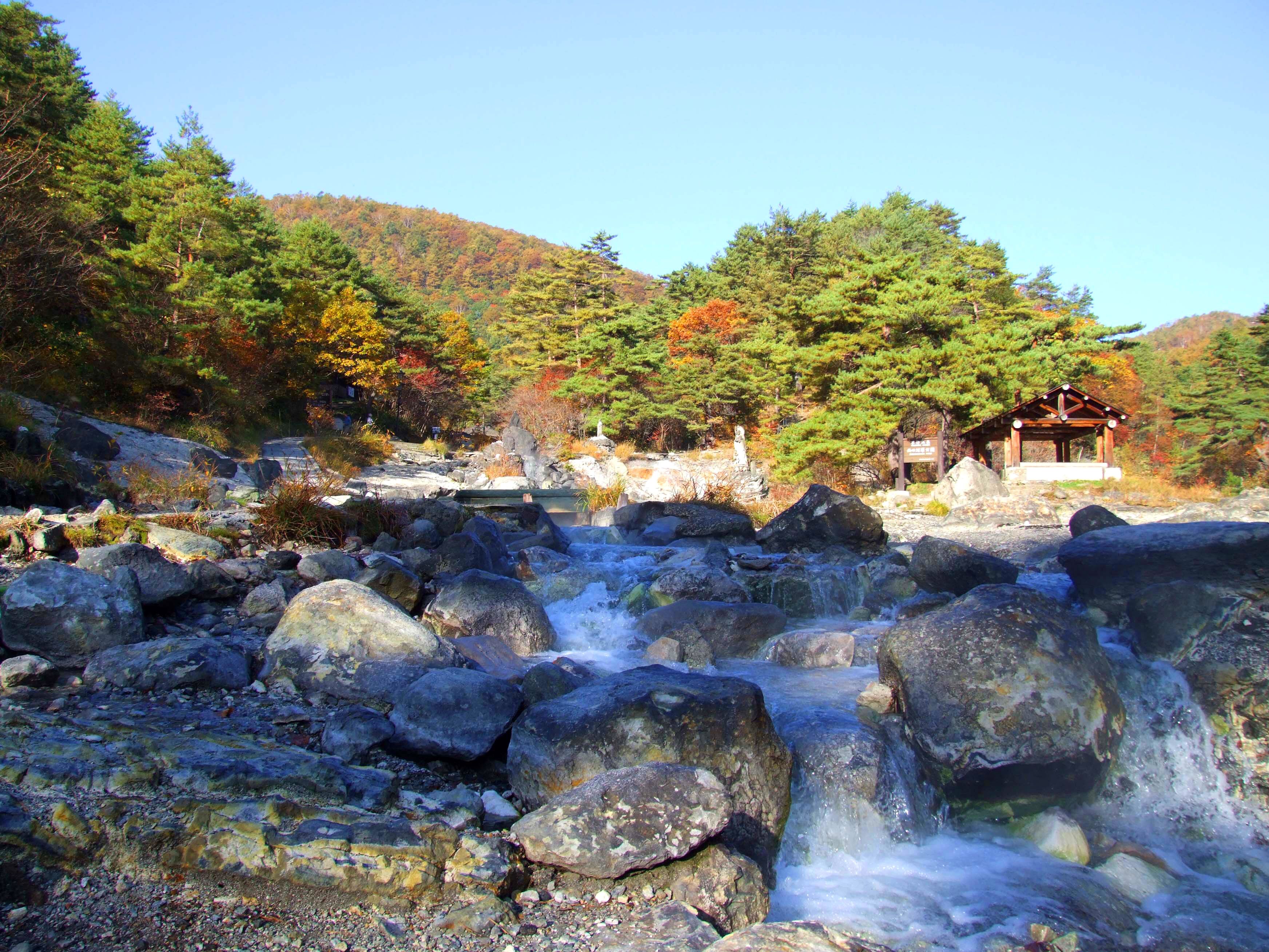 A natural landscape with a rocky stream, a wooden structure, and a forested hill.