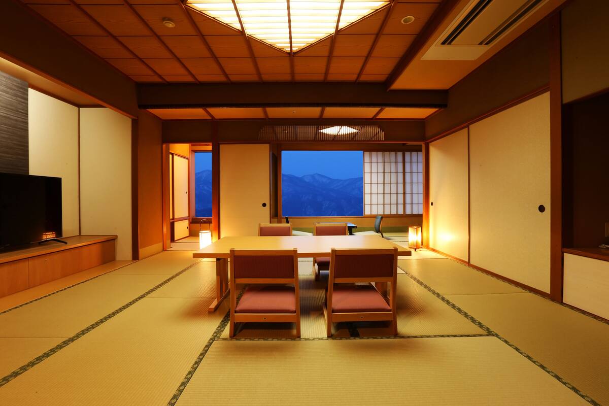 Une pièce de style japonais traditionnel, avec un sol recouvert de tatamis, une table basse entourée de chaises et un téléviseur placé à gauche.