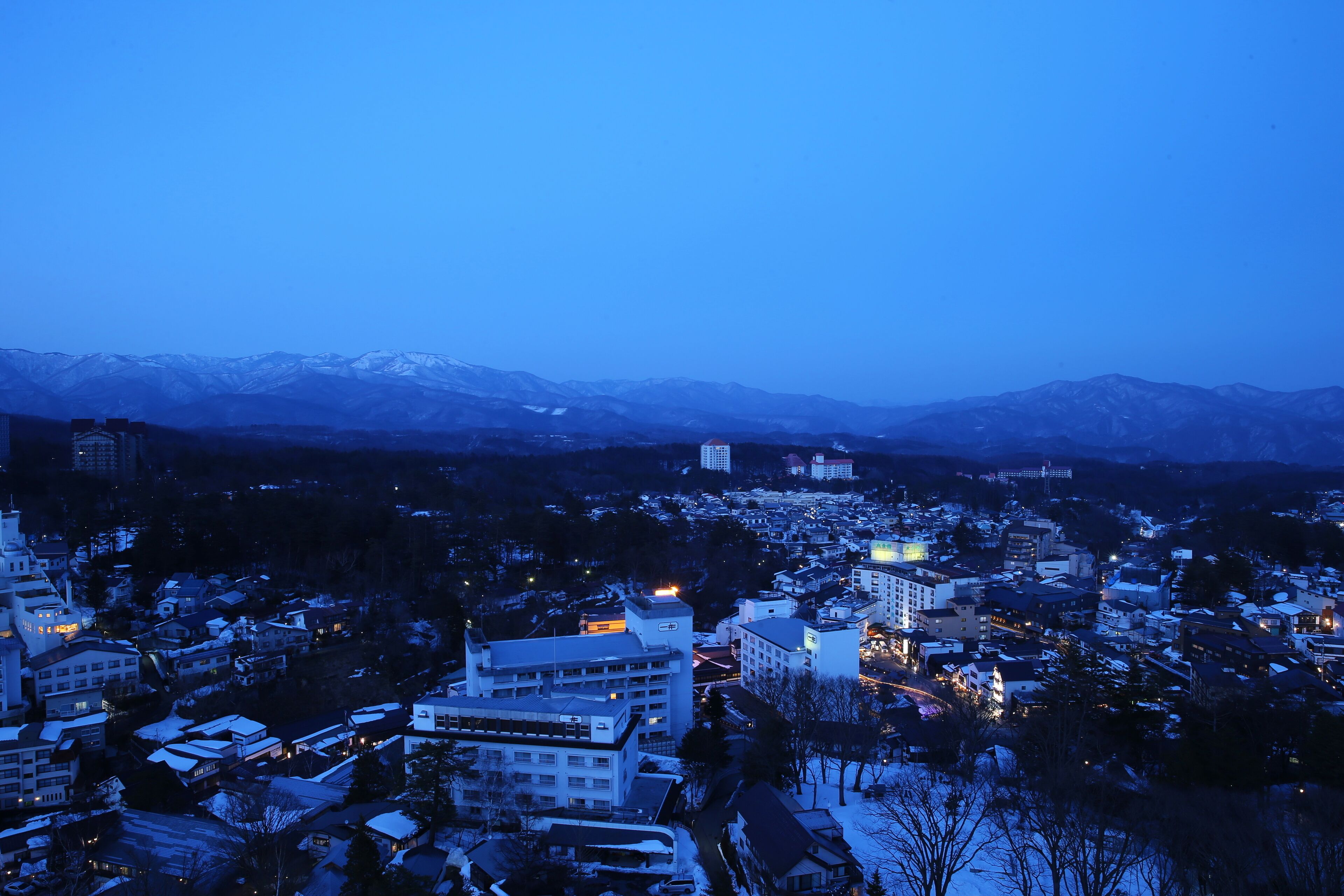 A cityscape at dusk with snow-covered buildings and mountains in the background.