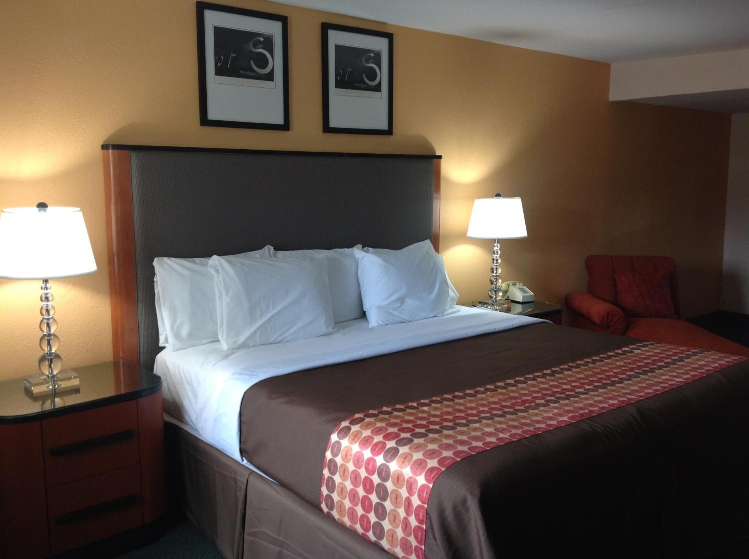 Une chambre d’hôtel avec un grand lit, deux tables de chevet avec des lampes, un fauteuil rouge et deux tableaux encadrés au mur.