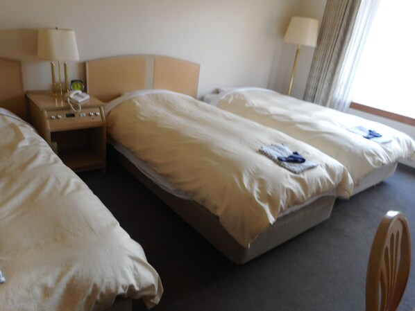 Quarto de hotel com duas camas de solteiro, mesa de cabeceira com abajur, telefone e janela com cortinas.