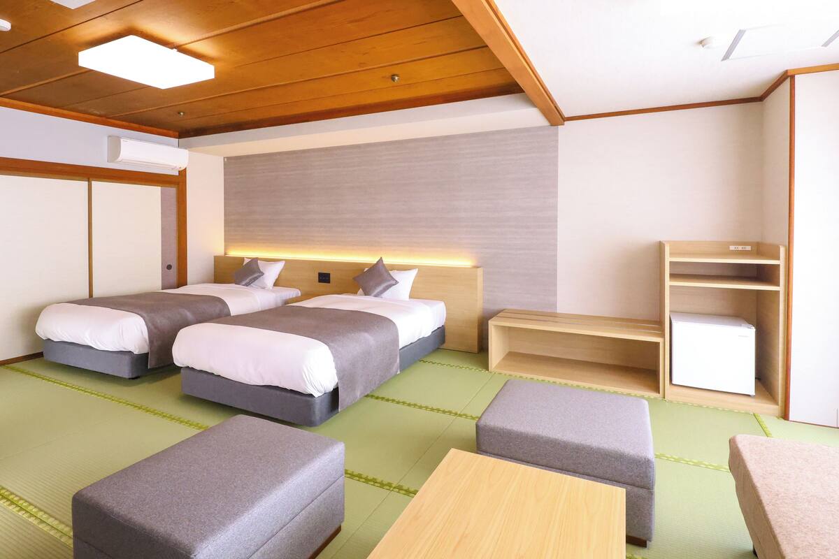 A hotel room with two beds, a small table, a bench, and a shelf.