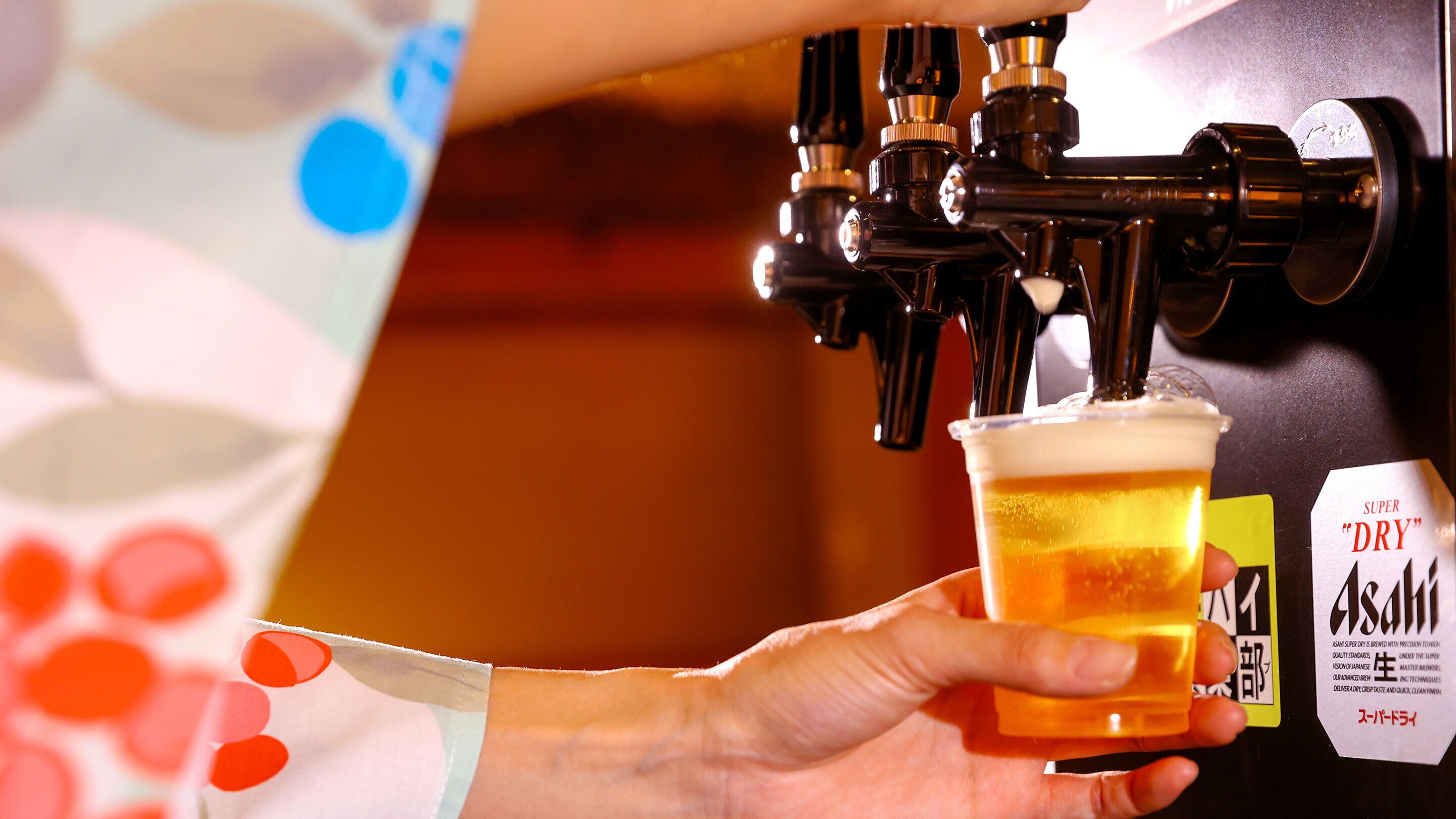 A hand holding a glass of beer with a label reading "SUPER DRY Asahi" being filled from a tap.