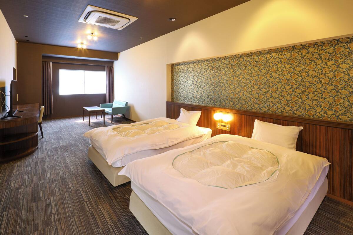 A hotel room with two beds, a desk, a chair, and a large window.