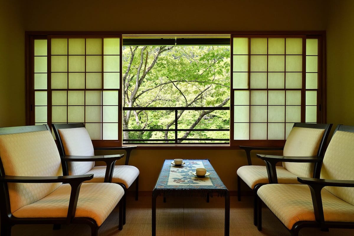 A traditional room with tatami flooring, a low table with a tea set, and a view of trees through shoji screens.