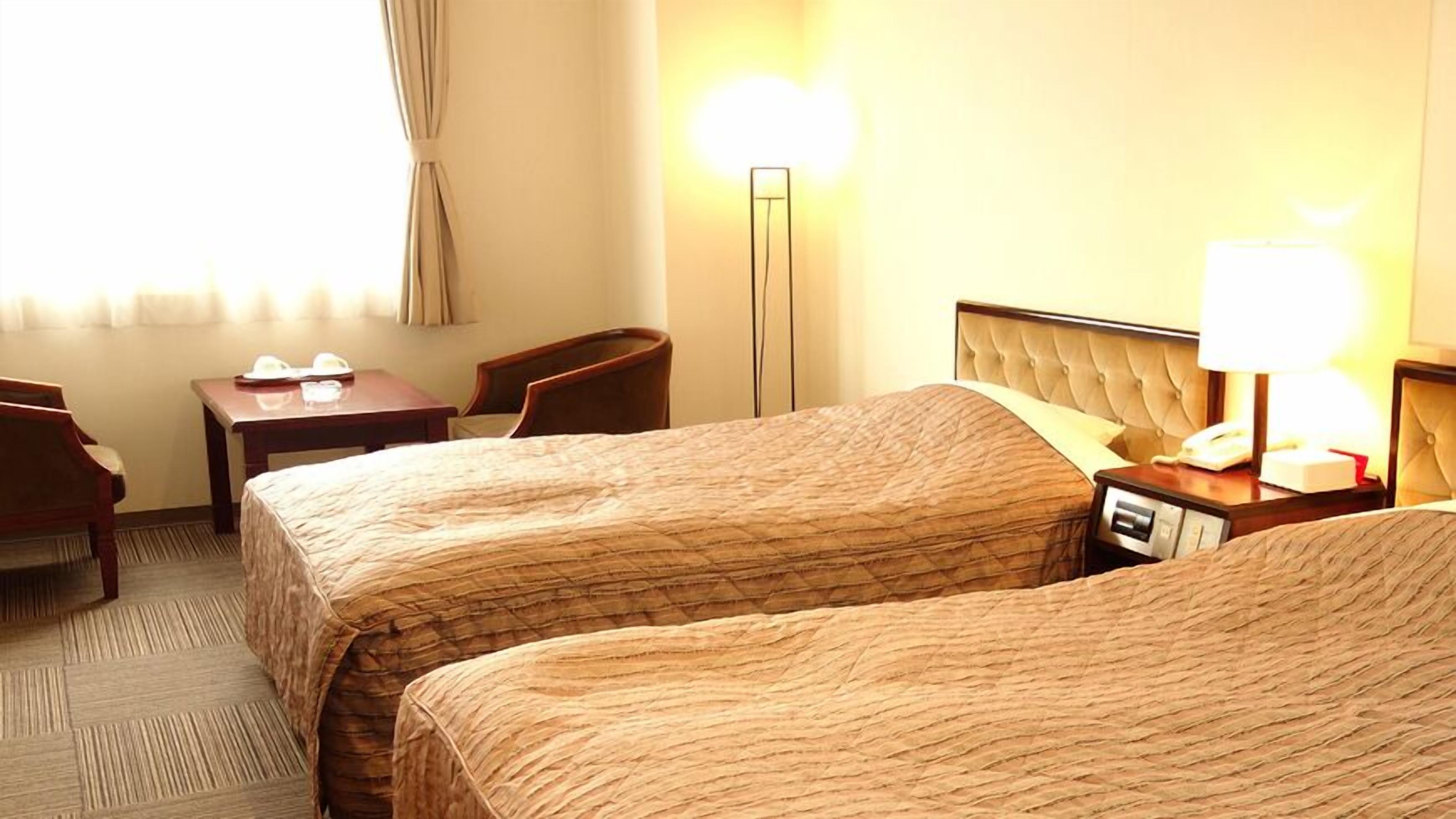 A hotel room with two beds, a desk, a chair, a lamp, and a bedside table.